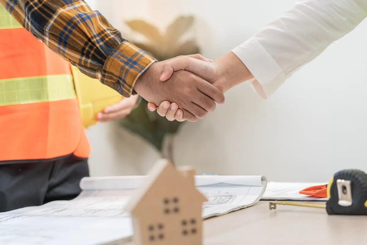 Apretón de manos sobre una mesa con planos y una pequeña casa de madera